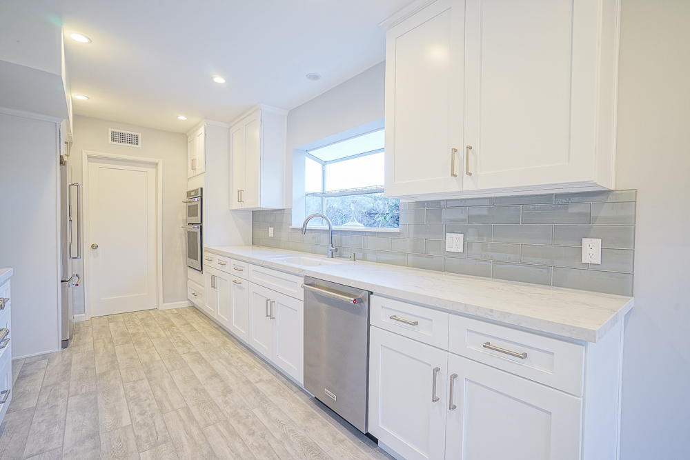 Kitchen from the far end showing a dishwasher, glass tile backsplash and a window over the sink